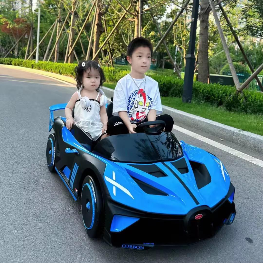 Two children in a blue toy car on a road with greenery in the background